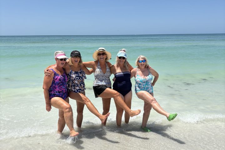 Five women in swimwear pose joyfully on a beach with clear water and blue sky.