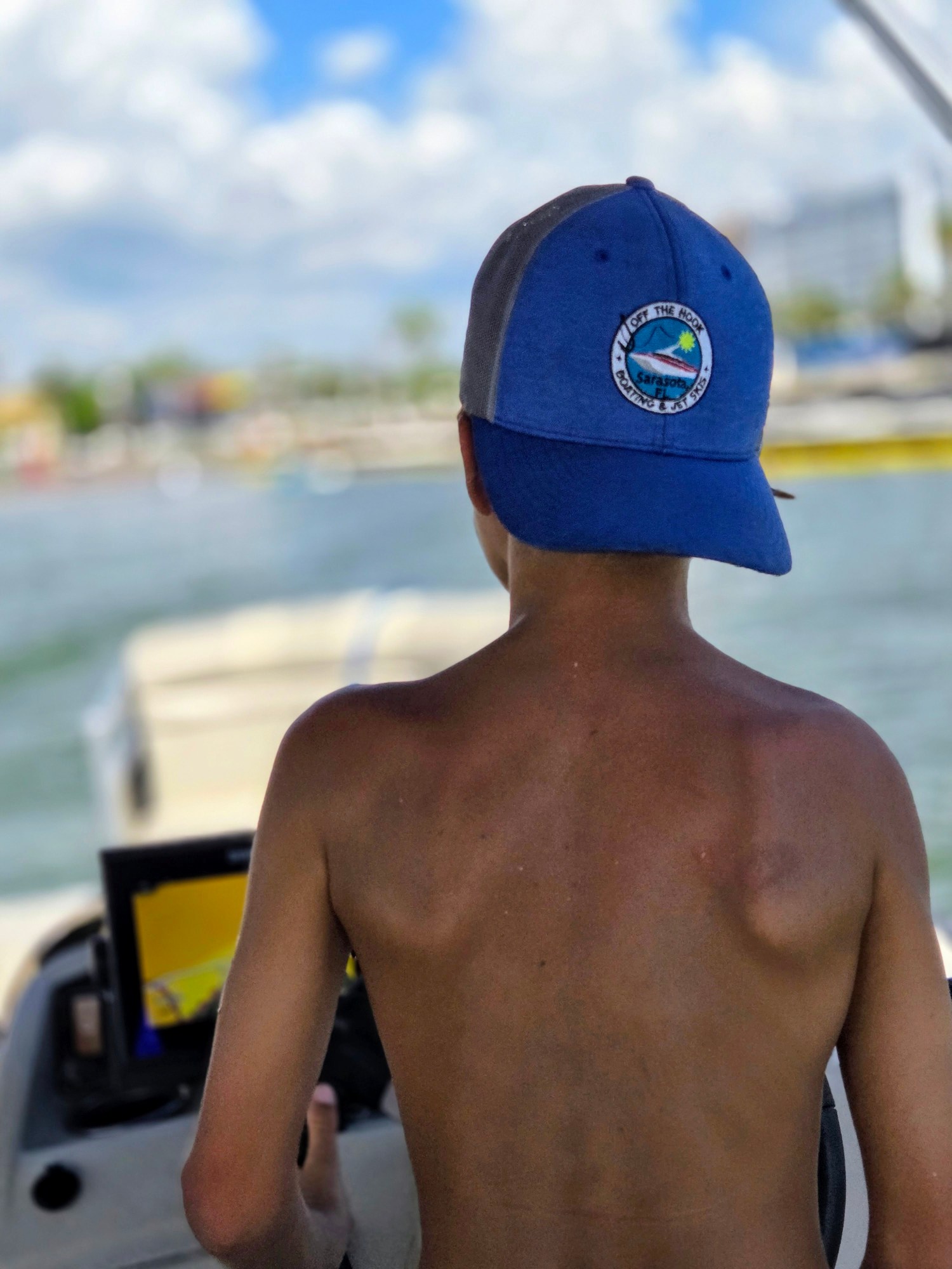 Shirtless child in blue cap steering a boat on a sunny day.